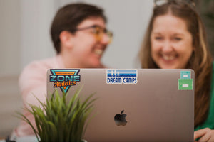 Laptop with stickers is in focus with two people smiling out of focus behind it