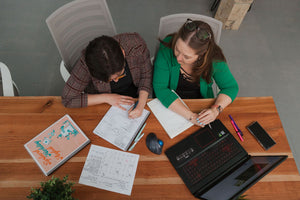 Two people sitting at a table surrounded by notebooks, pens, and a laptop